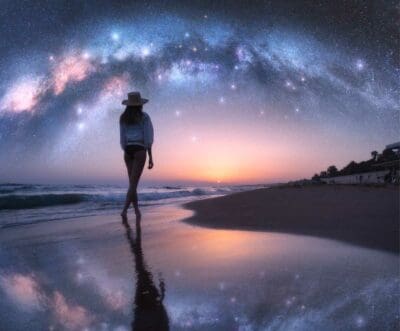Woman walks on beach at sunset. Stars in the sky are reflected by the water.